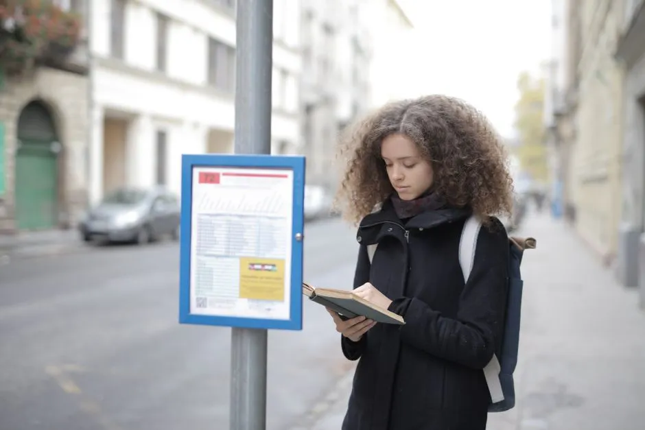 A student with a backpack waiting at a bus stop while reading, illustrative of student intercity bus travel